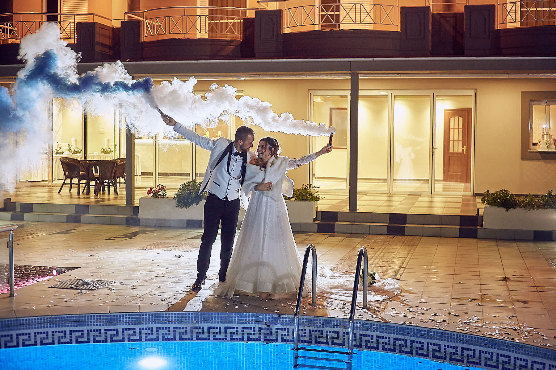 groom and bride holding smoke grenade, smoke around them by the hotel's pool